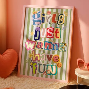 Puede incluir: Un estampado enmarcado con el texto "Girls Just Wanna Have Fun" en letras coloridas recortadas. El estampado está sobre un fondo a rayas verdes y blancas. También se ven una almohada en forma de corazón, una lámpara de lava y un tocadiscos.