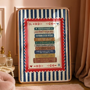 Puede incluir: Un cuadro enmarcado con una pila de libros, con títulos como "Heated Rivalry" e "I Love You". El marco dorado tiene un borde rojo y azul, y un fondo a rayas. El cuadro está apoyado contra una pared.