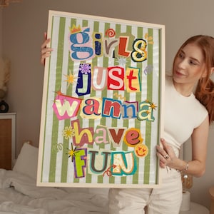May include: A framed art print with the text "Girls Just Wanna Have Fun" in colorful, cut-out style letters. The background is a green and white striped pattern. The frame is a light wood color.