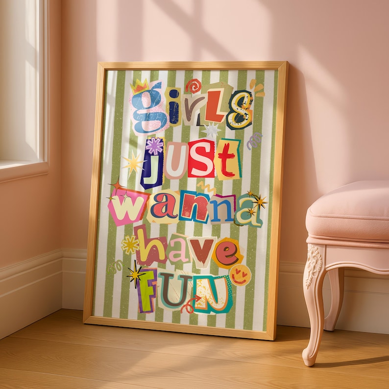 May include: A framed print with the text "Girls Just Wanna Have Fun" in colorful, cut-out style letters. The background features green and white vertical stripes. The frame is a light wood color.