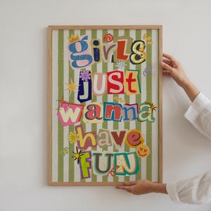 May include: A framed art print with the text "Girls Just Wanna Have Fun" in a colorful, cut-out style. The background features green and white vertical stripes. The frame is made of light-colored wood.