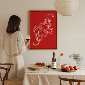Peut inclure: Une impression encadr&eacute;e rouge avec une illustration blanche de deux tigres se faisant face. L'impression est accroch&eacute;e &agrave; un mur blanc au-dessus d'une table avec une bouteille de vin, un verre de vin et un bol de fruits.