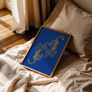 May include: A framed art print featuring two stylized gold tigers on a vibrant blue background. The tigers are depicted in a dynamic pose, set within a natural wood frame. The artwork is displayed on a bed with neutral-toned bedding.