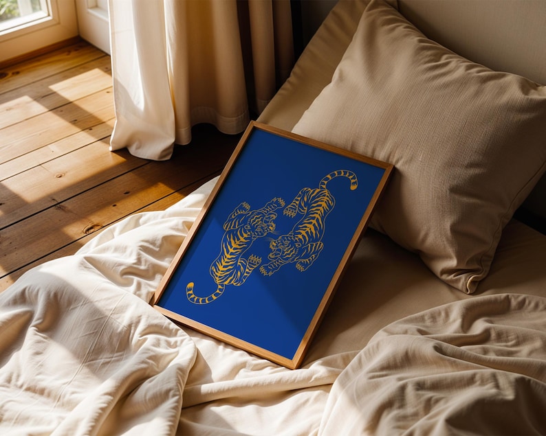 May include: A framed art print featuring two stylized gold tigers on a vibrant blue background. The tigers are depicted in a dynamic pose, set within a natural wood frame. The artwork is displayed on a bed with neutral-toned bedding.