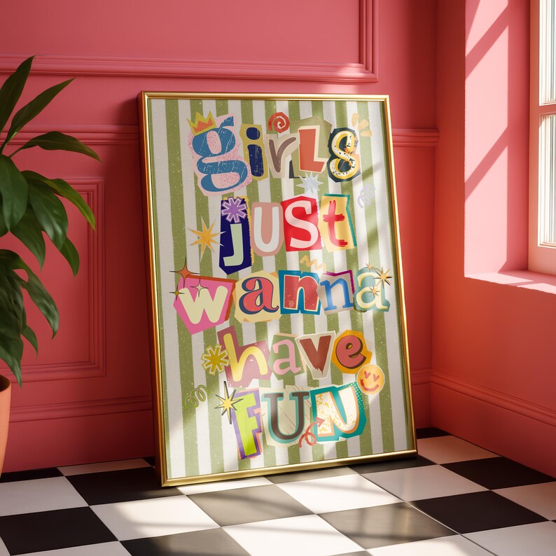May include: A framed art print with the text "Girls Just Wanna Have Fun" in colorful, cut-out style letters. The background features vertical green and white stripes. The frame is gold, and the artwork is displayed against a pink wall.
