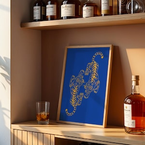 May include: A framed art print featuring two stylized yellow tigers on a vibrant blue background. The artwork is displayed on a wooden shelf, alongside a glass of amber liquid and a bottle of liquor.