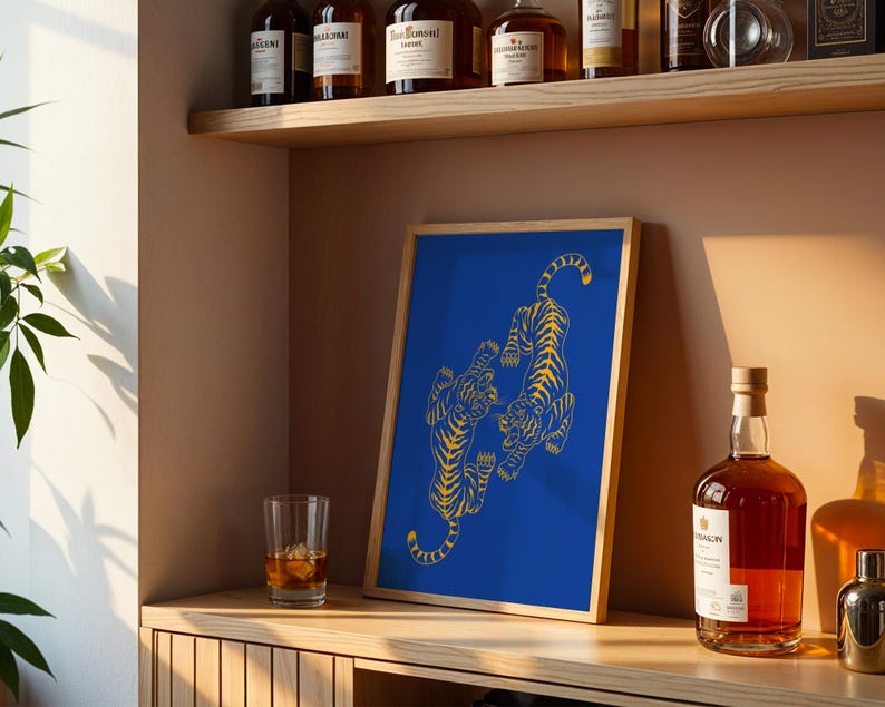 May include: A framed art print featuring two stylized yellow tigers on a vibrant blue background. The artwork is displayed on a wooden shelf, alongside a glass of amber liquid and a bottle of liquor.