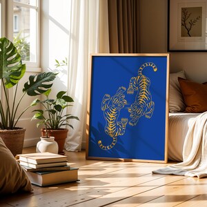May include: A framed art print featuring two stylized tigers in a yellow outline on a vibrant blue background. The artwork is displayed in a room with natural light, plants, and a bed with pillows and a blanket. The frame is a light brown color.
