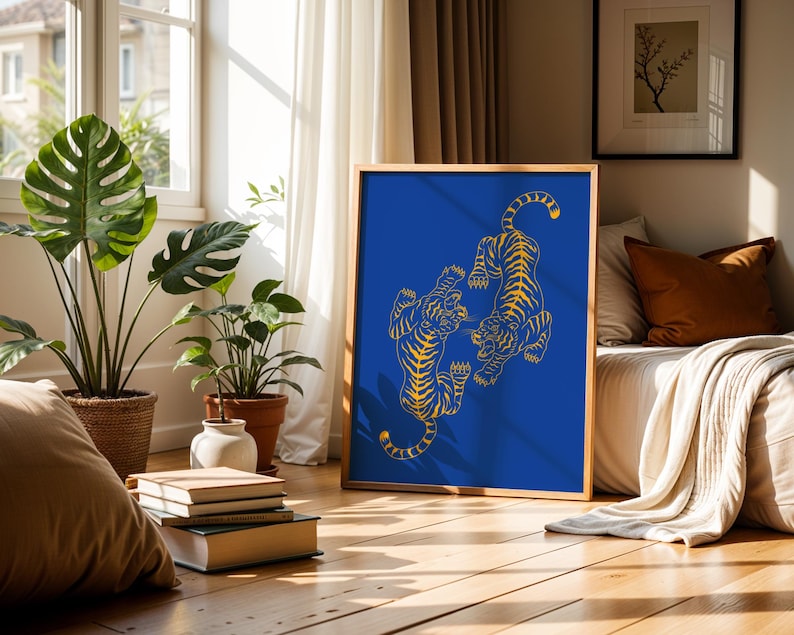 May include: A framed art print featuring two stylized tigers in a yellow outline on a vibrant blue background. The artwork is displayed in a room with natural light, plants, and a bed with pillows and a blanket. The frame is a light brown color.