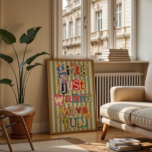 May include: A framed art print with the text "Girls Just Wanna Have Fun" in colorful, cut-out style letters. The print is set against a green and white striped background. The artwork is displayed in a living room setting with a sofa, chair, and potted plant.