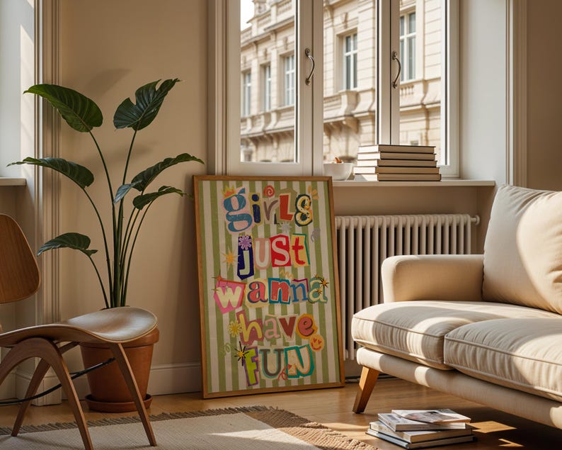 May include: A framed art print with the text "Girls Just Wanna Have Fun" in colorful, cut-out style letters. The print is set against a green and white striped background. The artwork is displayed in a living room setting with a sofa, chair, and potted plant.