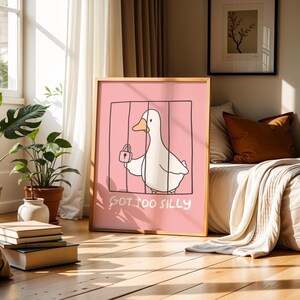 May include: A framed art print featuring a cartoon duck inside a cage, holding a padlock. The background is pink, and the text "GOT TOO SILLY" is written below the duck. The frame is made of wood.