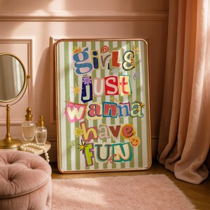May include: A framed art print with the text "Girls Just Wanna Have Fun" in colorful, cut-out style letters. The background is a green and white striped pattern. The frame has a gold finish.
