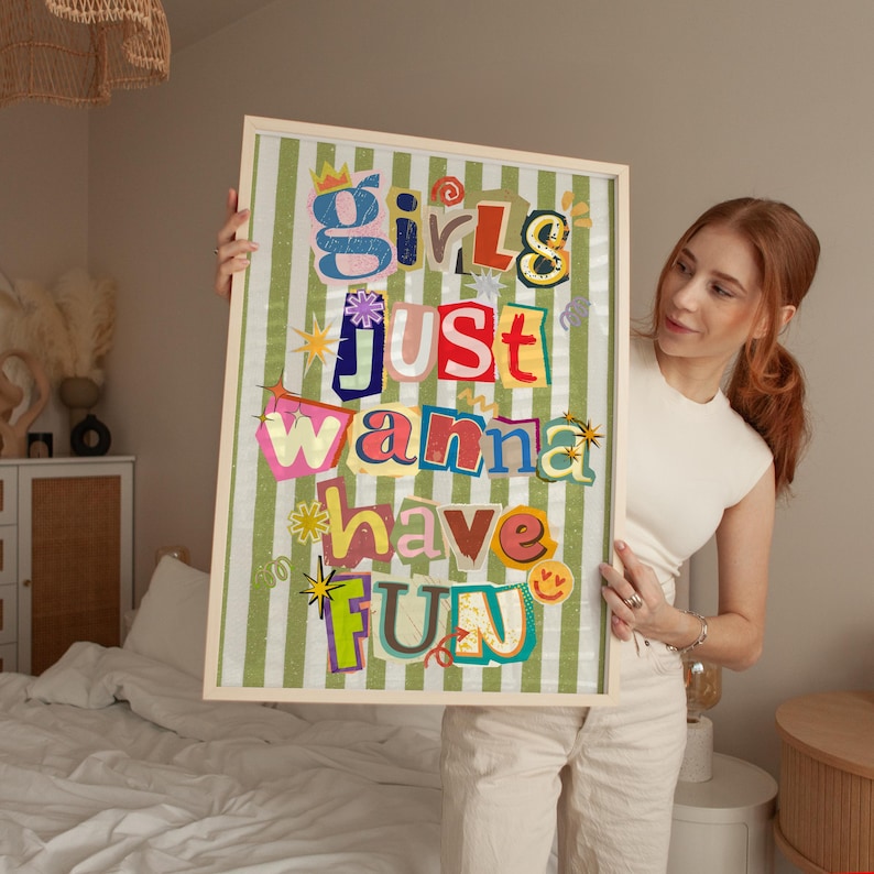 May include: A framed art print with the text "Girls Just Wanna Have Fun" in colorful, cut-out style letters. The background is a green and white striped pattern. The frame is a light wood color.