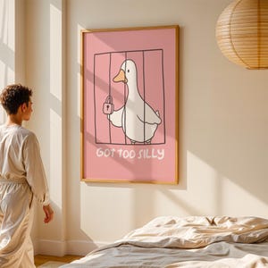 May include: A framed art print featuring a cartoon duck inside a cage, holding a padlock. The background is pink, and the text "GOT TOO SILLY" is below the duck. The frame is made of wood.