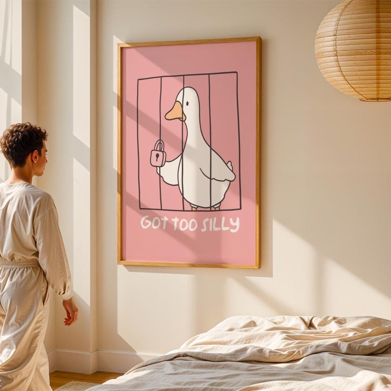 May include: A framed art print featuring a cartoon duck inside a cage, holding a padlock. The background is pink, and the text "GOT TOO SILLY" is below the duck. The frame is made of wood.