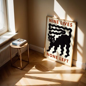 May include: A framed poster with a black cat illustration and the text "NINE LIVES NONE LEFT" in red. The poster is leaning against a wall in a room with a window and a small side table with magazines.