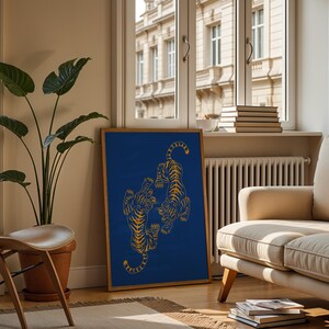 May include: A framed art print featuring two stylized tigers in gold against a deep blue background. The artwork is displayed in a living room setting, leaning against a wall near a window and a beige sofa. A large potted plant and a wooden chair are also visible.