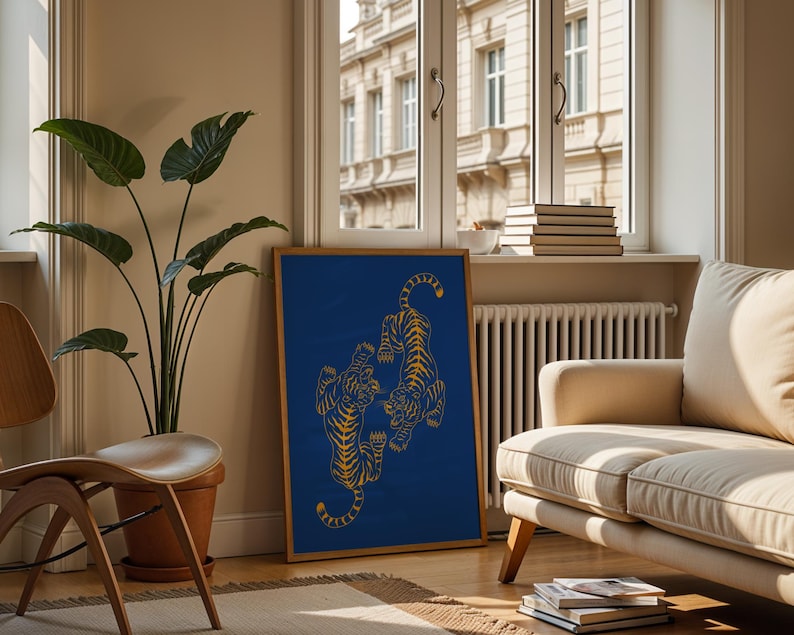 May include: A framed art print featuring two stylized tigers in gold against a deep blue background. The artwork is displayed in a living room setting, leaning against a wall near a window and a beige sofa. A large potted plant and a wooden chair are also visible.