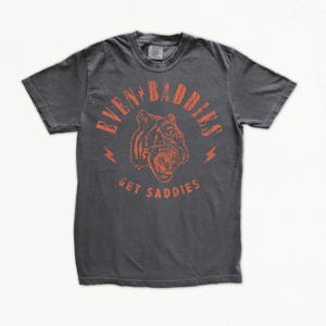 Può includere: T-shirt grigio scuro con grafica di tigre arancione e la scritta "EVEN BABIES GET SADDIES". Il design include fulmini. La maglietta ha girocollo e maniche corte.