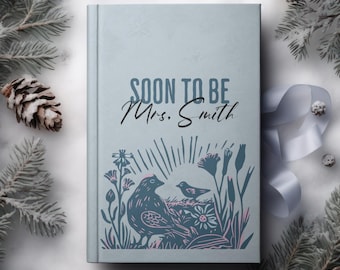 May include: A light blue journal with the text "SOON TO BE Mrs. Smith" in black script. The cover features a bird and floral design in blue and pink. The journal is surrounded by frosted pine branches and a ribbon.
