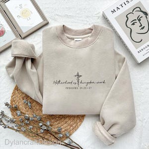 Puede incluir: Sudadera beige con el texto "Motherhood is kingdom work" y un dise&ntilde;o de cruz. El texto "PROVERBIOS 31:25-27" est&aacute; debajo. La sudadera est&aacute; doblada sobre una estera tejida con elementos decorativos.