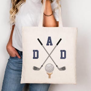 May include: Cream-colored tote bag featuring the word "DAD" in navy blue block letters, with crossed golf clubs and a golf ball on a tee. The bag is held by a person wearing a white shirt and blue jeans.