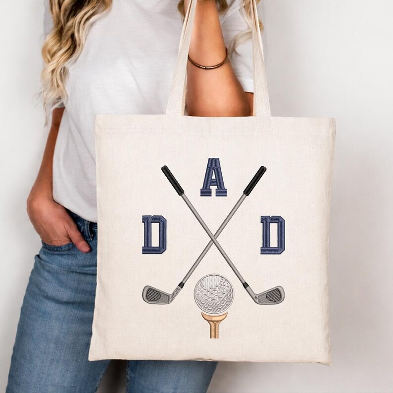 May include: Cream-colored tote bag featuring the word "DAD" in navy blue block letters, with crossed golf clubs and a golf ball on a tee. The bag is held by a person wearing a white shirt and blue jeans.