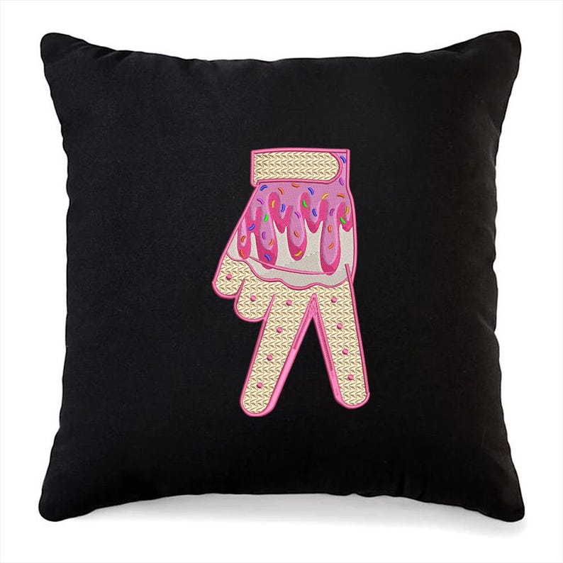 May include: Black throw pillow with a pink and white embroidered hand making the peace sign. The hand is decorated with pink frosting and colorful sprinkles.
