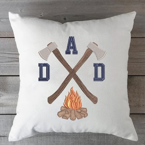 May include: White square pillow with an embroidered design. The design features two crossed axes, the word "DAD" in navy blue, and a campfire. The axes have brown handles and silver blades. The campfire has orange and yellow flames.