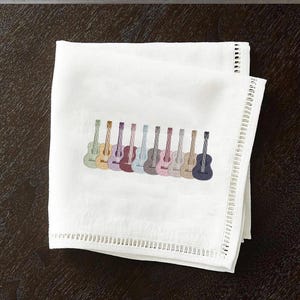 May include: White cloth napkin with a row of embroidered guitars in various colors. The guitars are in shades of green, yellow, purple, blue, and gray. The napkin has a decorative border.
