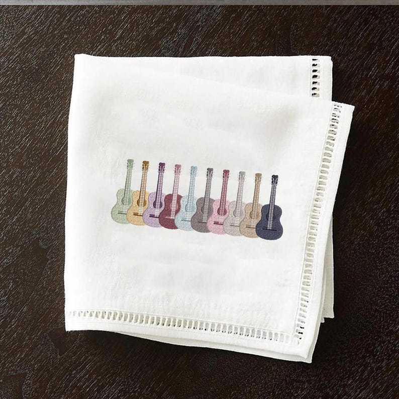 May include: White cloth napkin with a row of embroidered guitars in various colors. The guitars are in shades of green, yellow, purple, blue, and gray. The napkin has a decorative border.