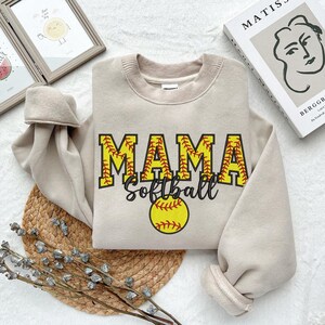 May include: A beige sweatshirt with the text "MAMA Softball" in black and red lettering. A yellow softball with red stitching is below the text.