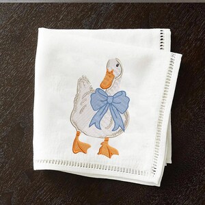 May include: White embroidered kitchen towel with a blue bow and a white duck with an orange beak.