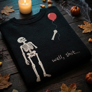 May include: A black crewneck sweater featuring an embroidered white skeleton figure. The skeleton is positioned below a red balloon held by a detached skeletal hand. The text "well, shit..." is embroidered in white script on the lower right side.