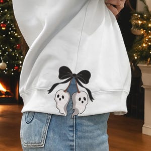 Può includere: Felpa bianca con un disegno ricamato di due figure a forma di fantasma appese a un fiocco nero. I fantasmi hanno piccoli occhi scuri. La felpa è abbinata a jeans blu.