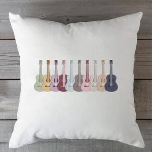 May include: White square pillow featuring an embroidered design of ten colorful guitars. The guitars are arranged in a row, each with a different pastel color, including green, yellow, purple, and blue. The pillow is made of a soft, white fabric.