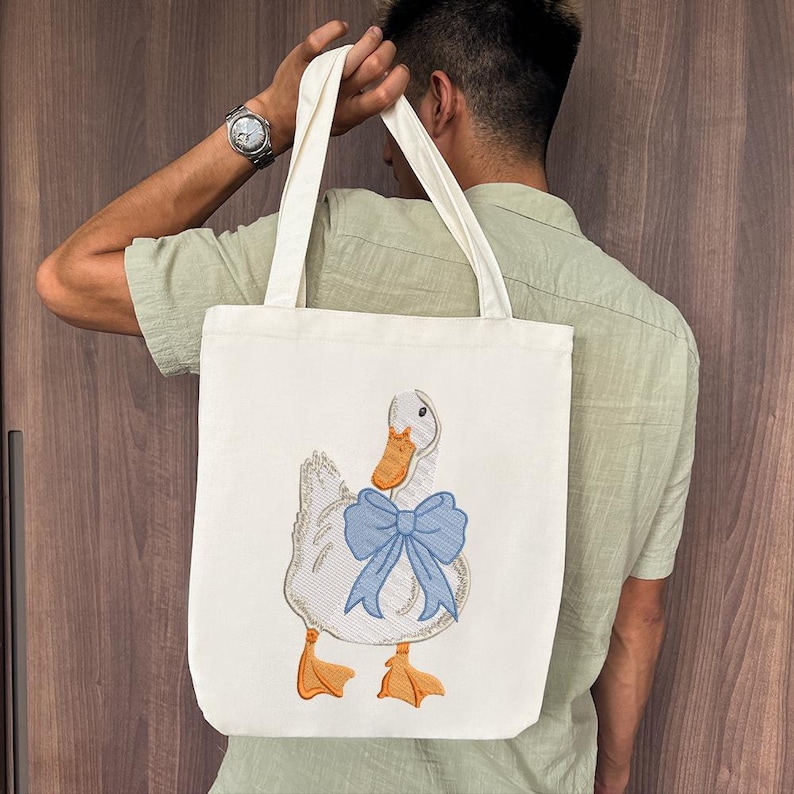 May include: A white canvas tote bag with a blue bow and an embroidered image of a white duck with an orange beak.