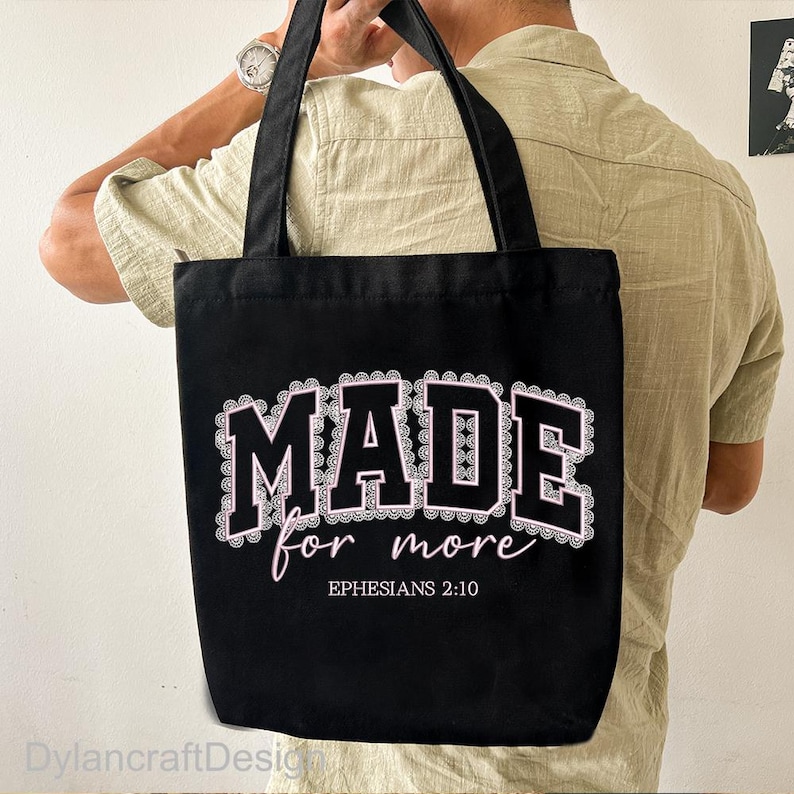 May include: Black tote bag featuring the text "MADE for more" in white and pink, with "Ephesians 2:10" below. The bag has black handles and is held by a person. The design is framed by a decorative border.