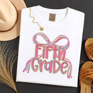 May include: White t-shirt with a colorful embroidered design that says "Fifth Grade" in pink and blue. A gold necklace and a tan hat are next to the shirt. The shirt has a tag that says "KORBY'S BOUTIQUE BY IVYKE".