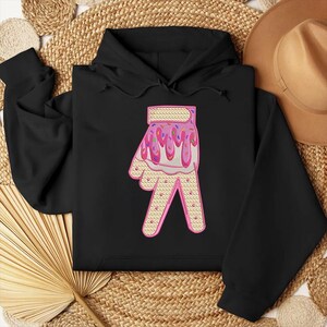 May include: Black hoodie with a pink and white hand design. The hand is covered in pink frosting and sprinkles.