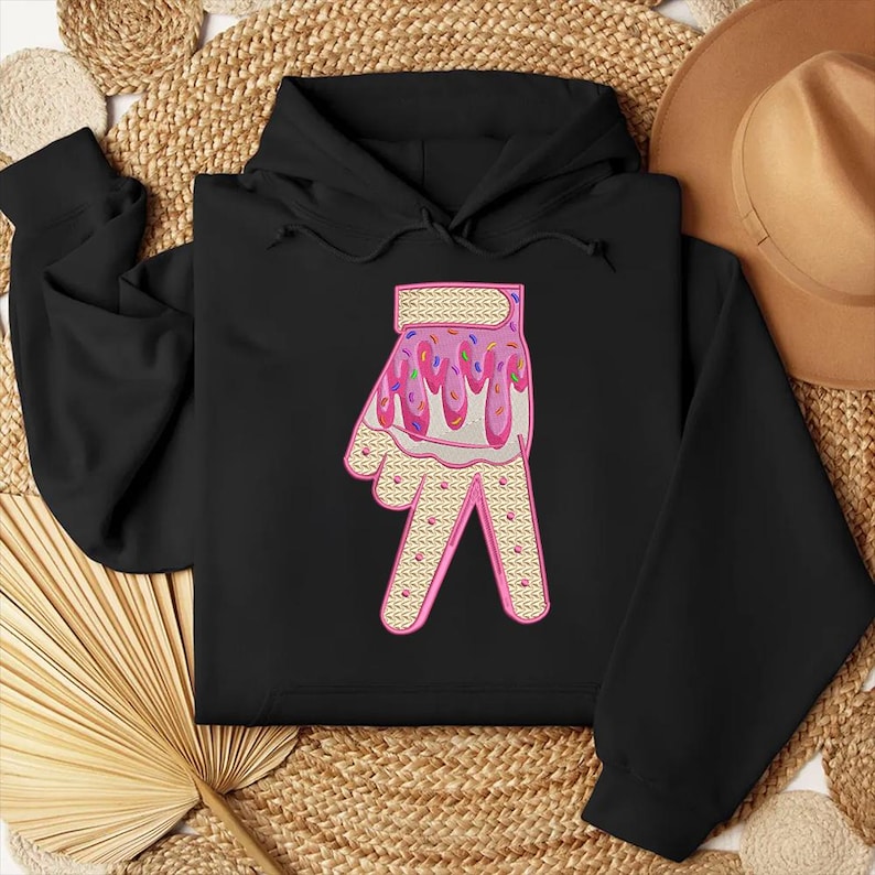 May include: Black hoodie with a pink and white hand design. The hand is covered in pink frosting and sprinkles.