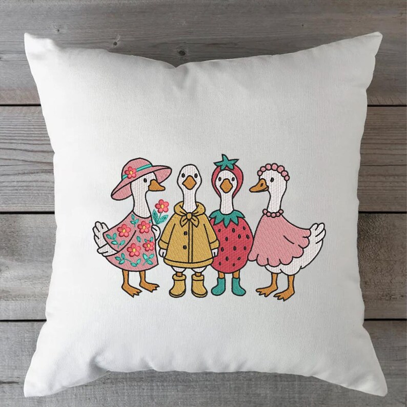 K&ouml;nnte beinhalten: Vier gestickte G&auml;nse, die verschiedene Outfits tragen. Eine Gans tr&auml;gt ein rosa Kleid und einen Hut, eine andere tr&auml;gt einen gelben Regenmantel, eine weitere tr&auml;gt ein Erdbeerkost&uuml;m und die letzte tr&auml;gt ein rosa Kleid mit einer Perlenkette.