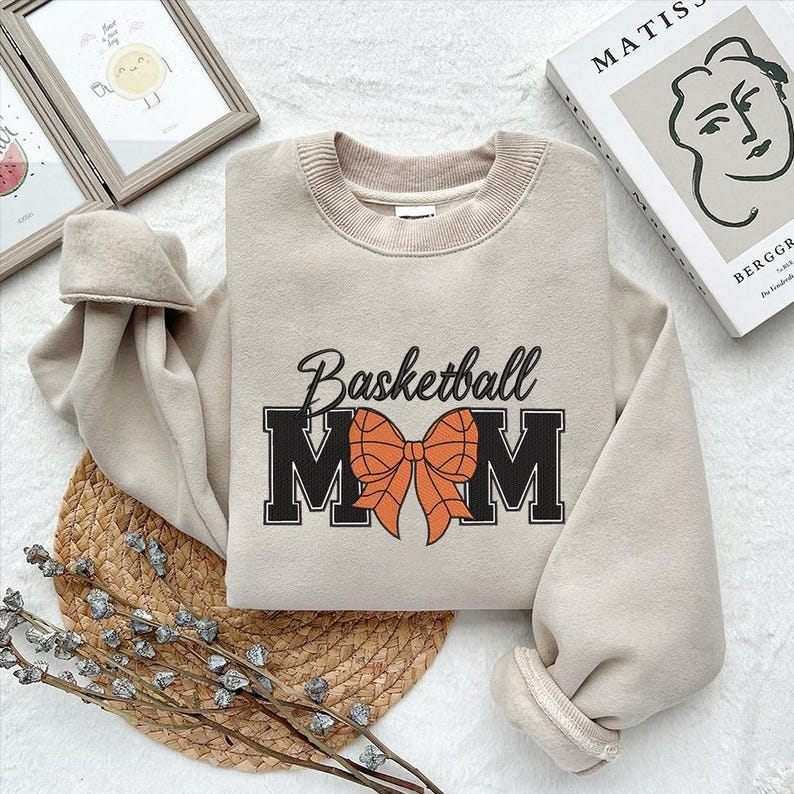 Peut inclure: Un sweat-shirt beige avec le texte "Basketball Mom" en noir et orange. Le mot "Mom" est en grandes lettres avec un grand n&oelig;ud orange au milieu.