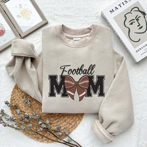 Peut inclure: Un sweat-shirt beige avec le texte "Football Mom" en lettres noires avec un n&oelig;ud en forme de ballon de football marron.