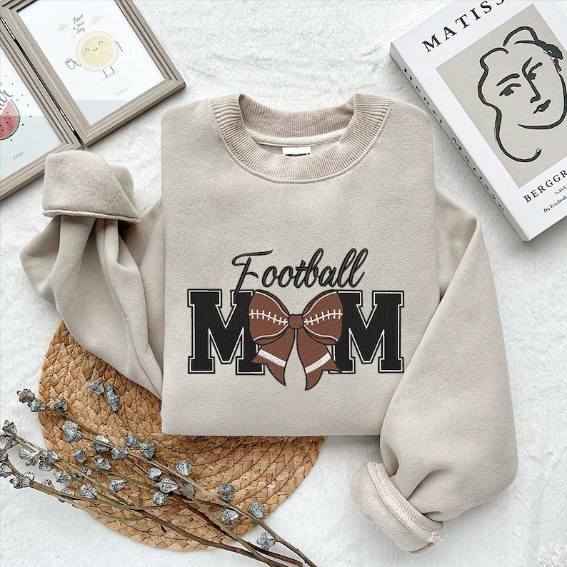 Peut inclure: Un sweat-shirt beige avec le texte "Football Mom" en lettres noires avec un n&oelig;ud en forme de ballon de football marron.