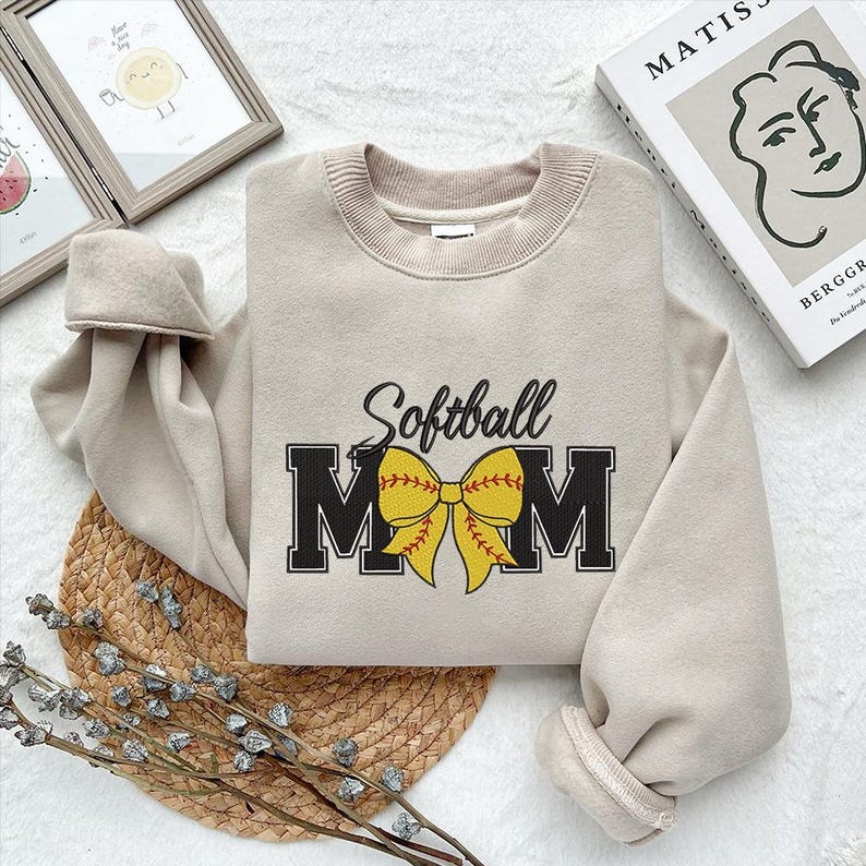 Peut inclure: Un sweat-shirt beige avec le texte "Softball Mom" en noir avec un n&oelig;ud de softball jaune et rouge.