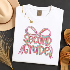 May include: White t-shirt with a colorful bow design and the words "Second Grade" in a playful font. A gold necklace and a tan hat are visible in the background. The shirt is likely for a student.