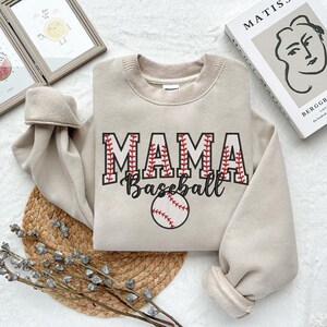 May include: A beige sweatshirt with the text "MAMA Baseball" in red and white stitching. A baseball with red stitching is centered below the text.