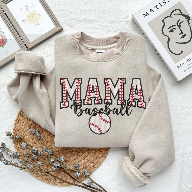May include: A beige sweatshirt with the text "MAMA Baseball" in red and white stitching. A baseball with red stitching is centered below the text.